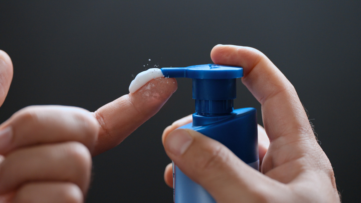 Close-up of hands dispensing white lotion from a blue pump bottle onto a finger, with a dark, blurred background.