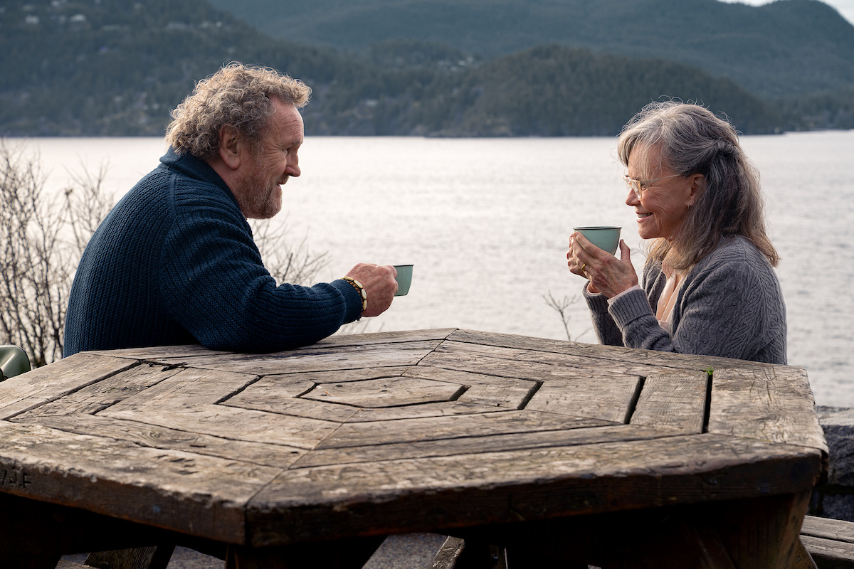 Colm Meaney as Ethan and Sally Field as Tova sit across from each other at a rustic wooden picnic table outdoors, smiling and enjoying hot drinks with a serene lake and forested mountains in the background.