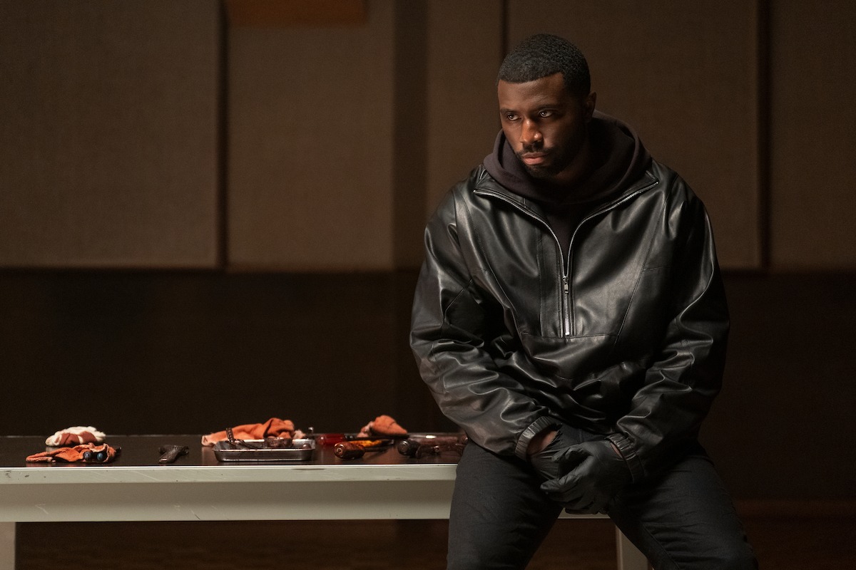A man in a black leather jacket sits on the edge of a white table indoors, with gloves on, looking serious. Assorted objects and food are spread on the table beside him. The room has soundproofed panels on the wall.