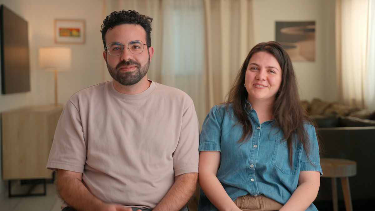 Two people sitting side by side in a cozy, well-lit living room with neutral decor, wall art, a lamp, curtains, and a sofa visible in the background, both facing the camera and smiling.