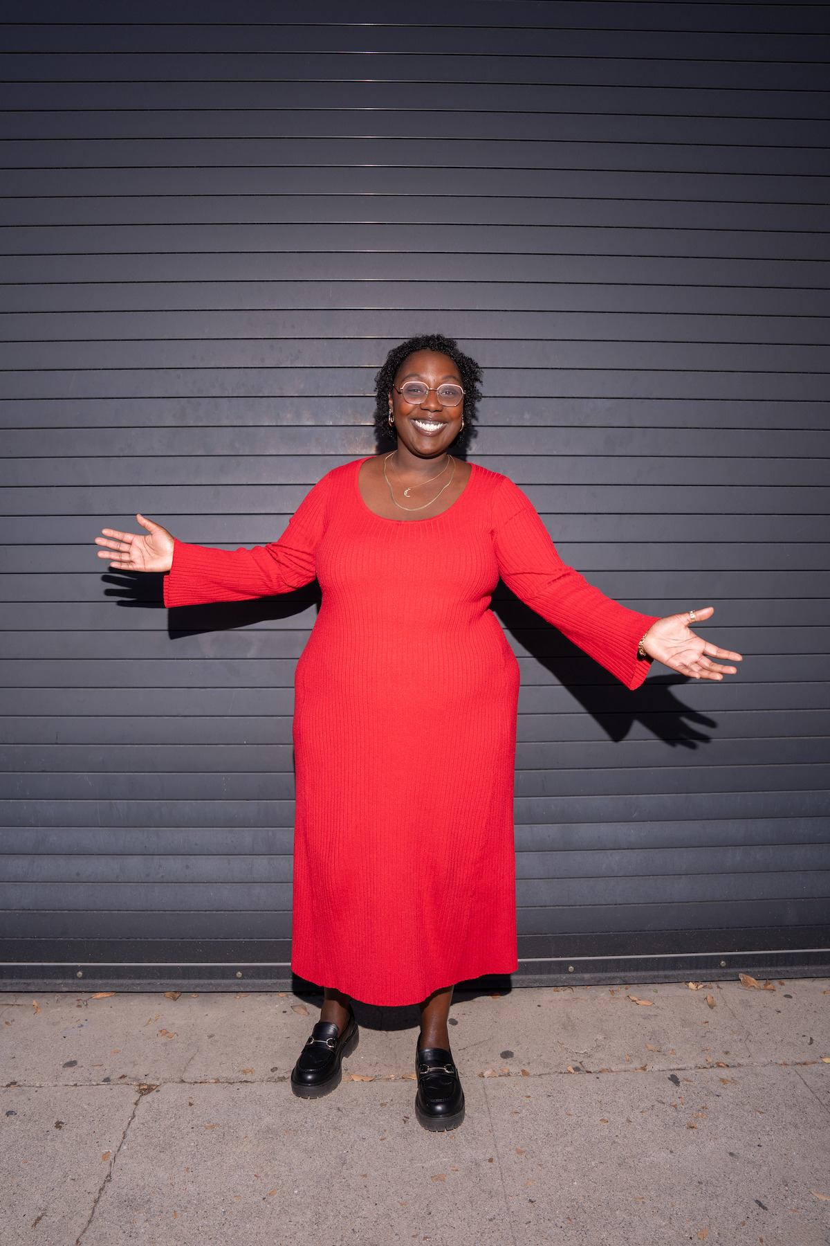 Person in a bright red dress stands smiling with arms open wide in front of a dark metal garage door, on a concrete sidewalk, during the day.