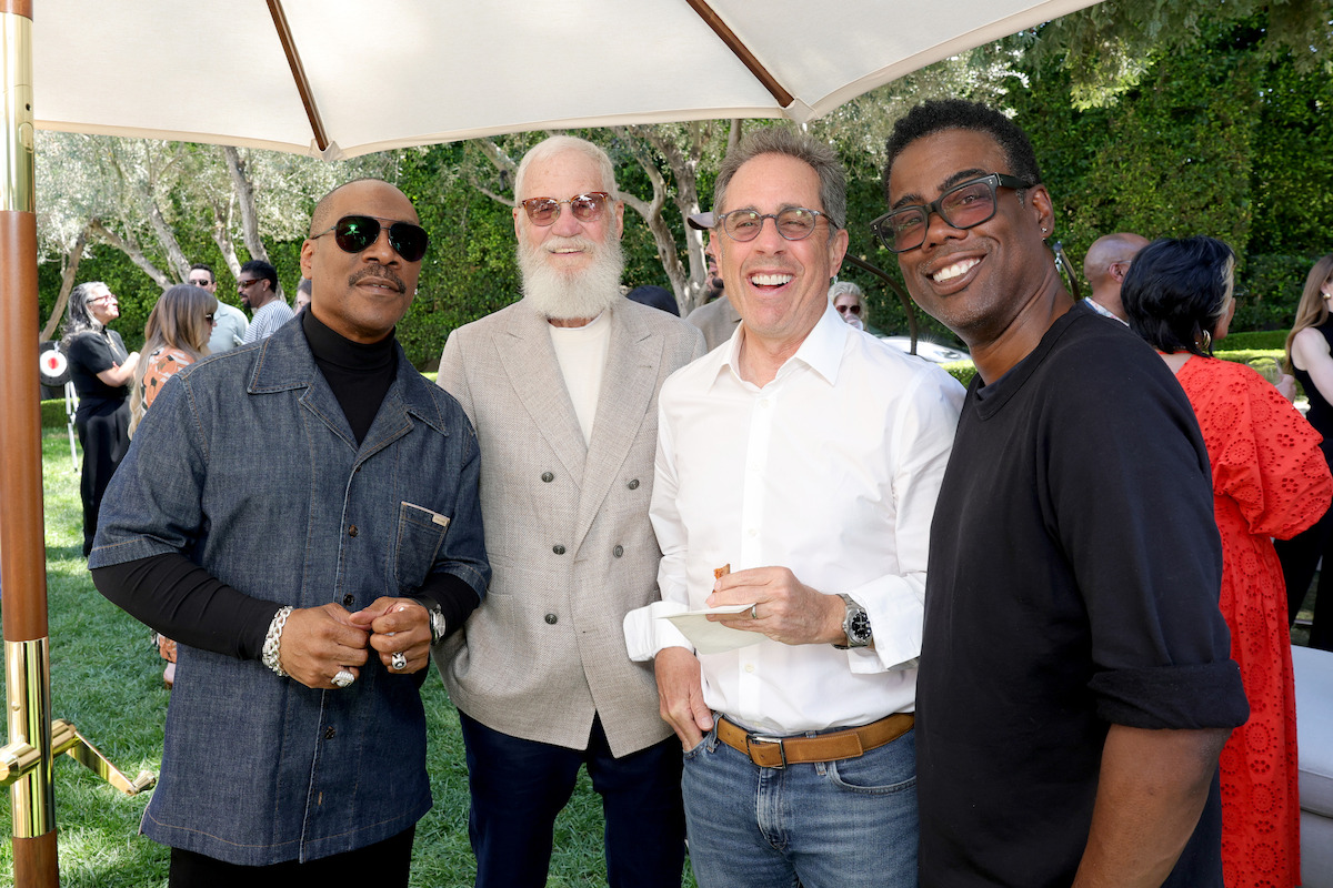 Four men smiling and posing together outdoors at a daytime social gathering under a large patio umbrella, surrounded by greenery and other people in the background.