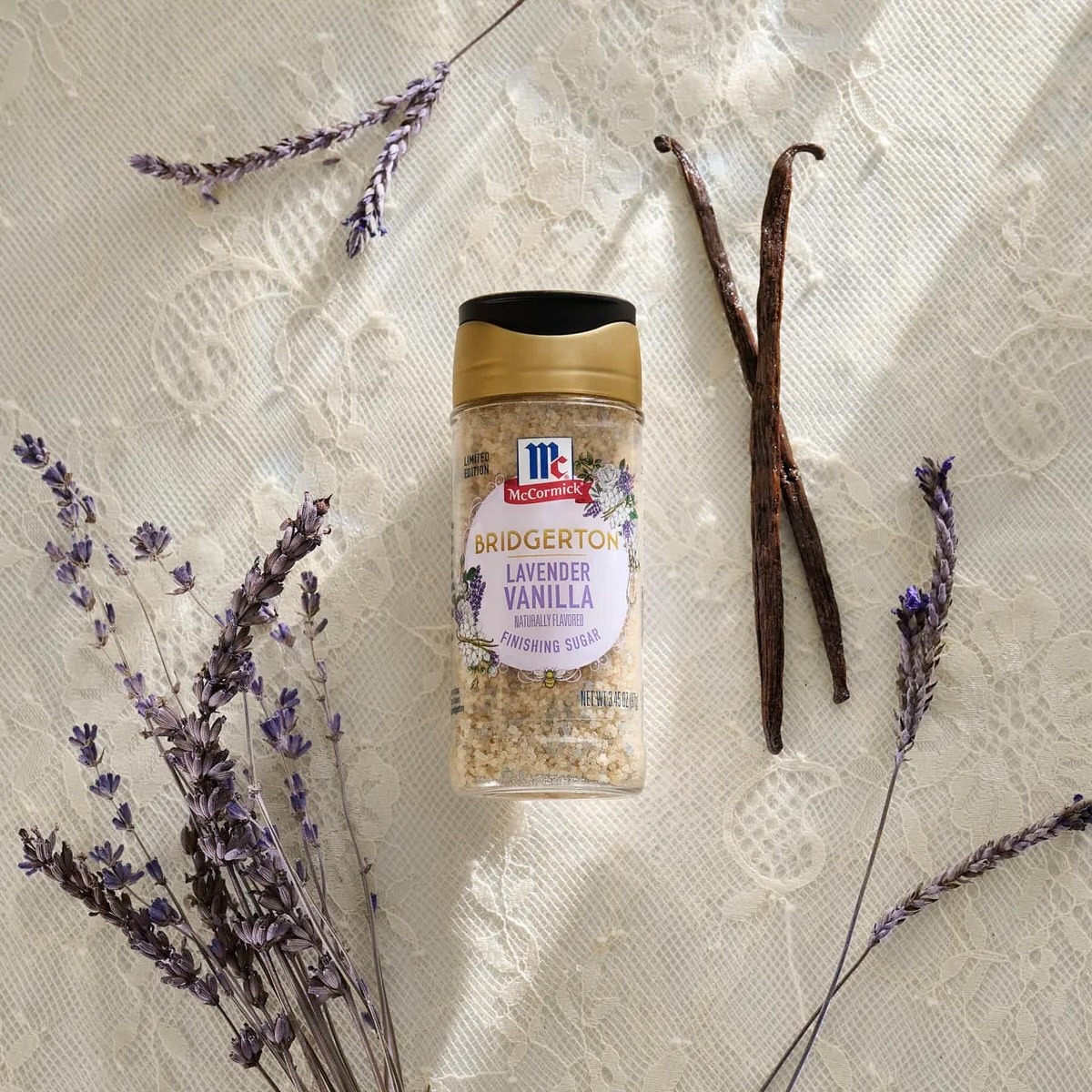 A jar of lavender vanilla sugar lies on lace fabric, surrounded by dried lavender flowers and vanilla beans, in soft natural light.