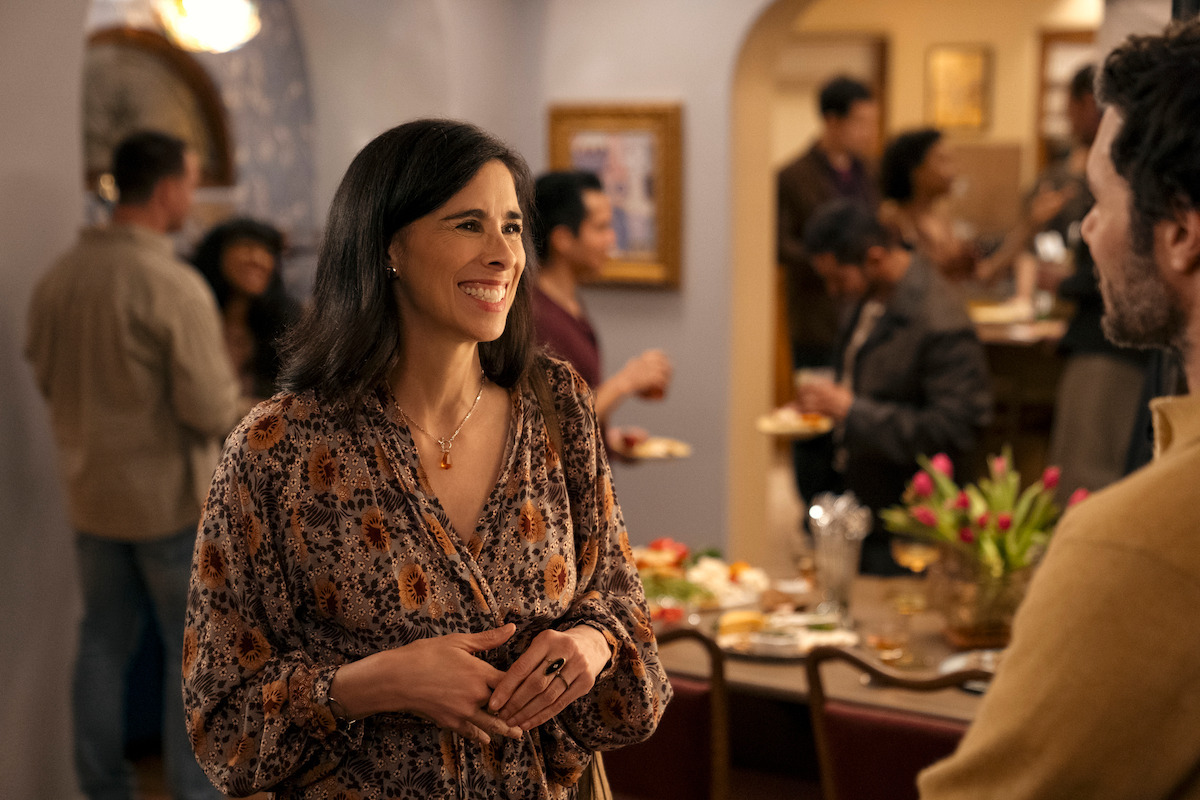 Woman smiling and talking to a man at a lively indoor gathering, with people socializing and food on a table in a warmly lit room decorated with flowers and paintings.