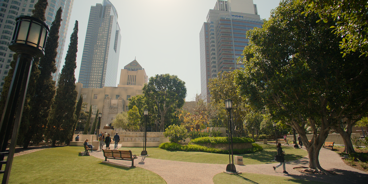 City park with green lawns, trees, benches, and walking paths, surrounded by tall modern buildings. People are walking and sitting, and sunlight filters through the trees, creating a peaceful urban oasis atmosphere.