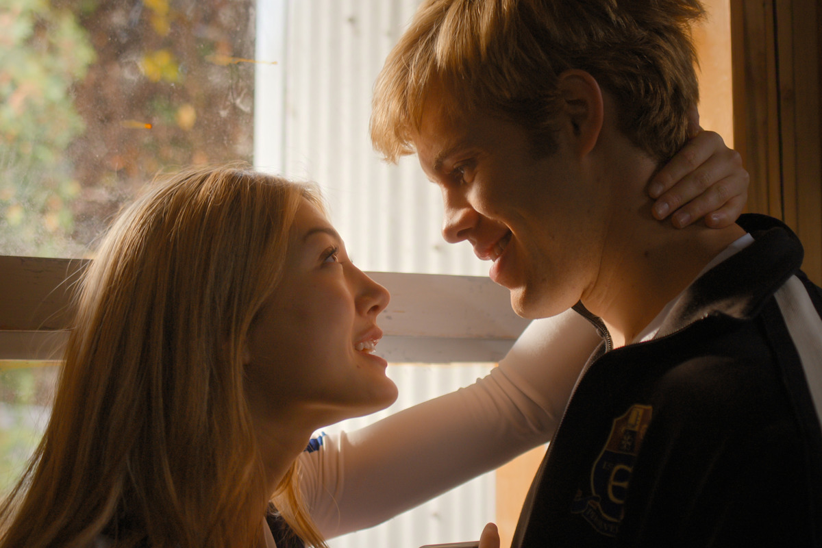 A young woman and man stand close indoors by a window, gazing into each other’s eyes and smiling warmly. The light is soft and natural, suggesting a tender or romantic moment in a cozy, sunlit setting.