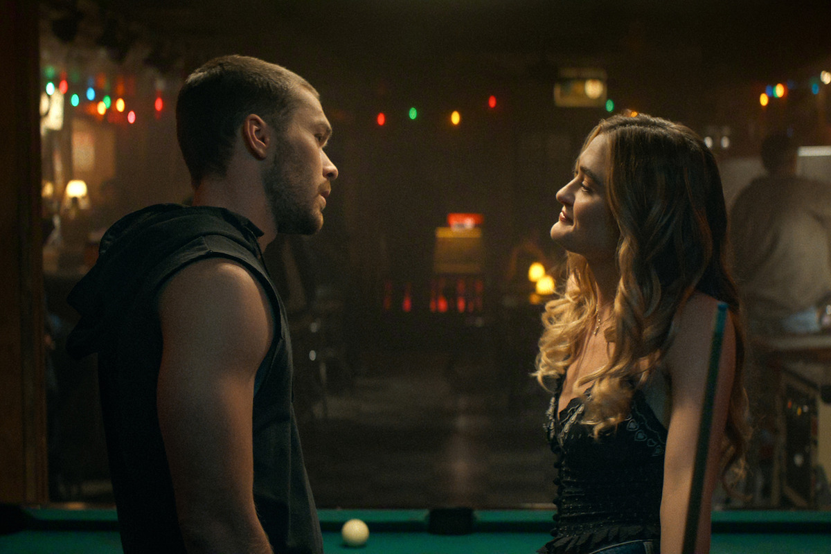 A man and woman stand close by a pool table in a dimly lit bar with string lights, facing each other and engaged in conversation, background out of focus and atmospheric.