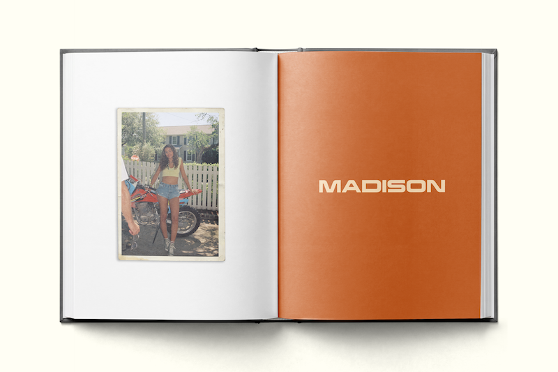 Open photo book showing a woman standing by a dirt bike in a yard with a white picket fence on the left page, and a bold orange page with the word "MADISON" on the right page.