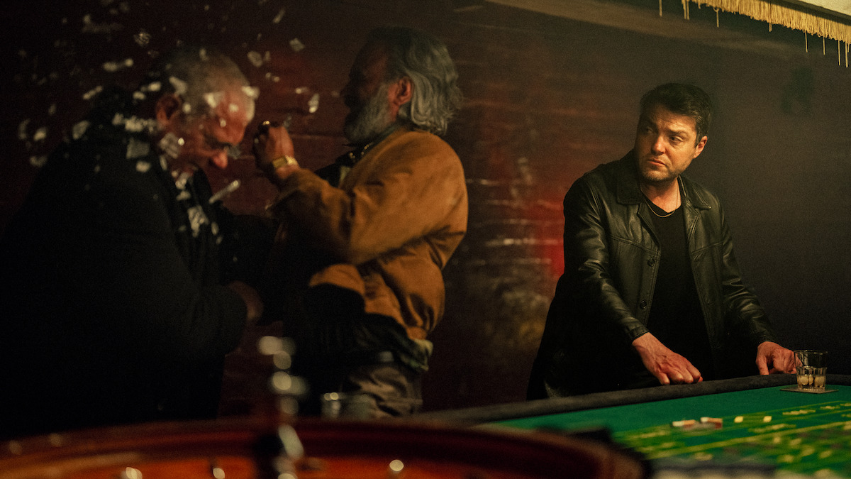 Three men in a dimly lit casino or gambling room; one man is hitting another, breaking glass, while a third man in a black jacket watches with a drink in hand at the game table.