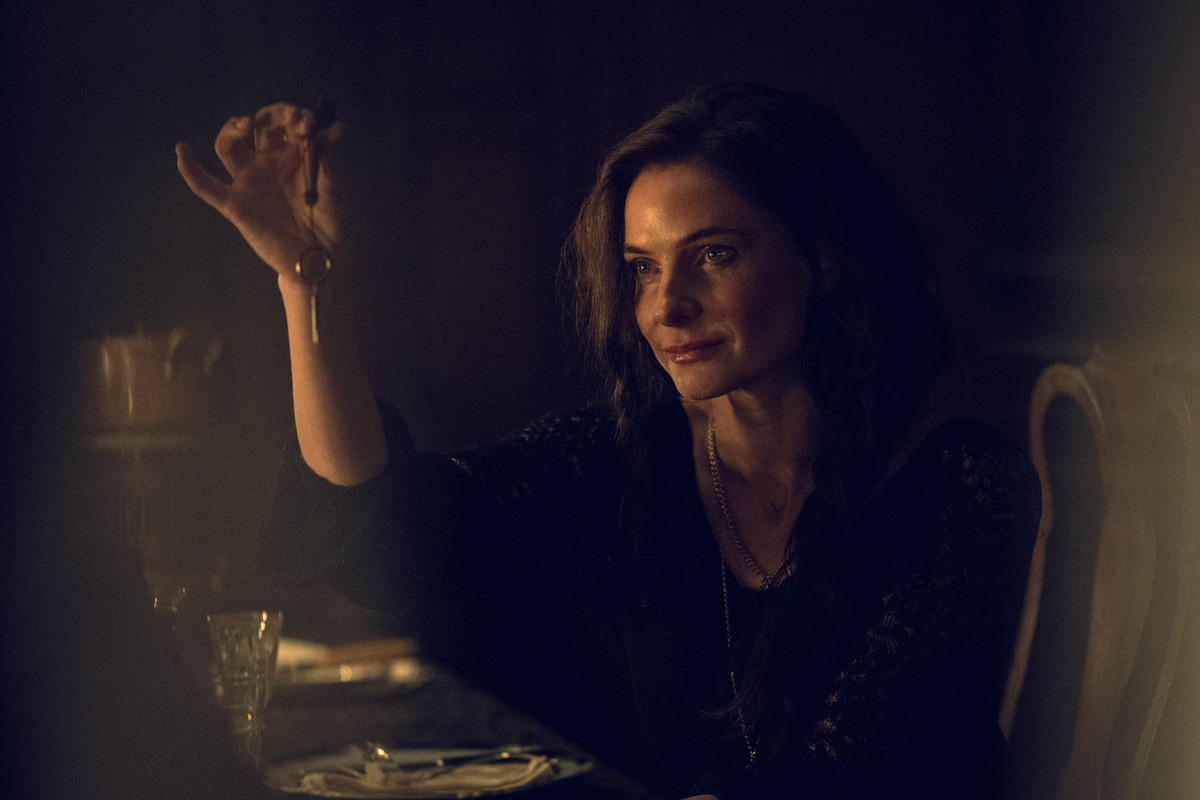 Woman sitting at a dimly lit dining table, holding up a set of keys with a serious expression, surrounded by a dark and moody atmosphere.