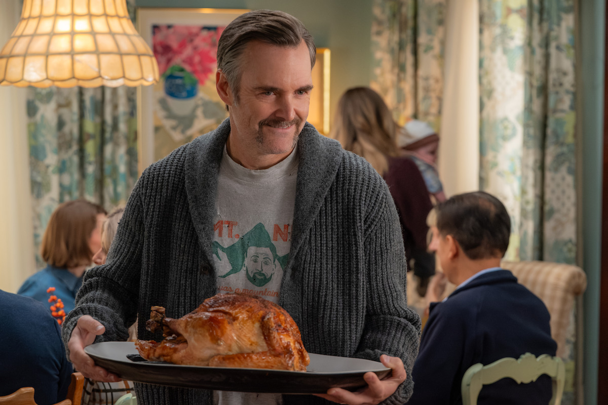 Man holding a roasted turkey on a tray, smiling in a cozy dining room filled with people, patterned curtains and warm lighting, suggesting a festive family meal or holiday gathering.