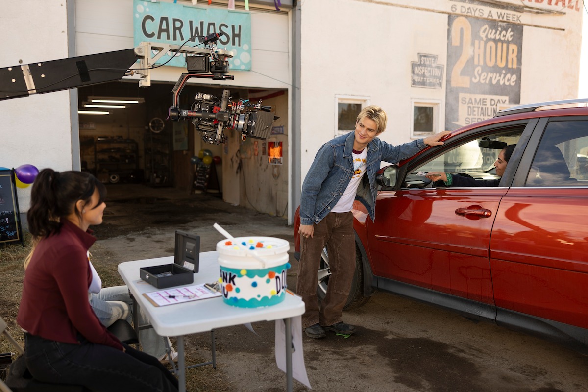 A film set at a car wash: a camera crane films a man talking to a woman in a red SUV, while another woman sits at a table. The environment is casual, with signs and garage doors visible.