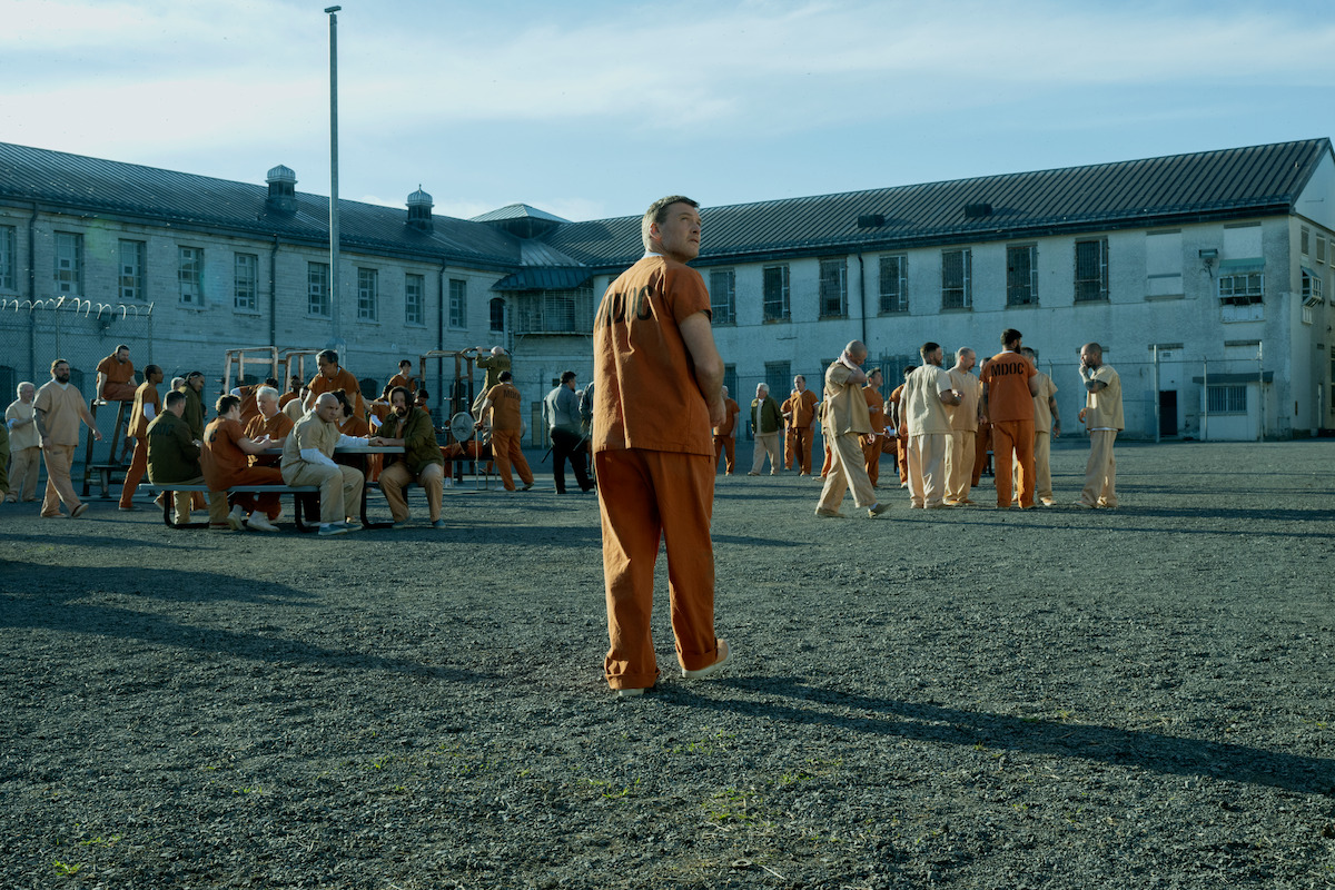 A group of inmates in orange uniforms gather and interact in the outdoor yard of a large prison complex under a blue sky, with one man standing in the foreground looking around.