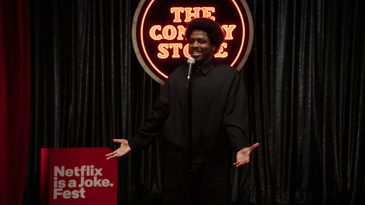 Stand-up comedian performing on stage at The Comedy Store with a microphone, smiling, in front of a red neon sign and dark curtains. Netflix is a Joke Fest sign is visible on the left.