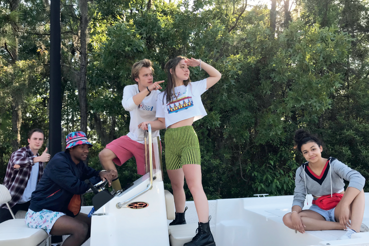 Five young people on a boat surrounded by trees, some sitting and some standing, enjoying a sunny day outdoors with relaxed expressions and casual summer outfits.