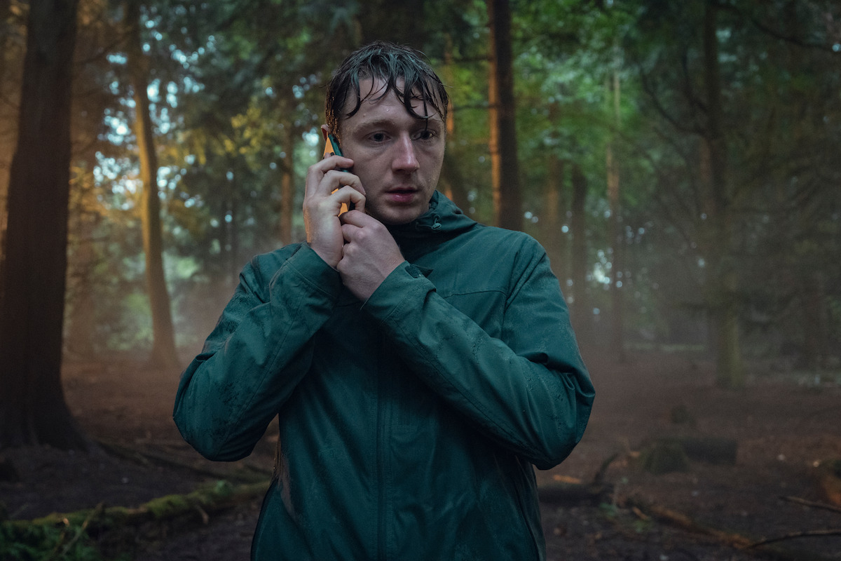A wet, worried person in a rain jacket stands in a misty, dense forest, holding a phone to their ear, surrounded by tall trees and fallen branches, with diffused sunlight in the background.