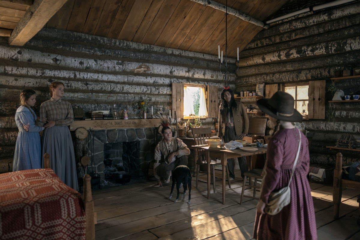 Five people in 19th-century clothing stand and sit in a rustic log cabin with wooden walls and ceiling, stone fireplace, simple furniture, and warm natural light from small windows. A dog sits near a man by the fireplace.