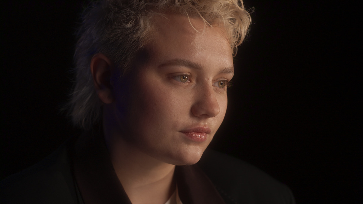 Person with short, light blonde hair in a dark jacket, looking pensive, is softly lit against a dark background in a close-up portrait, creating a calm and introspective atmosphere.