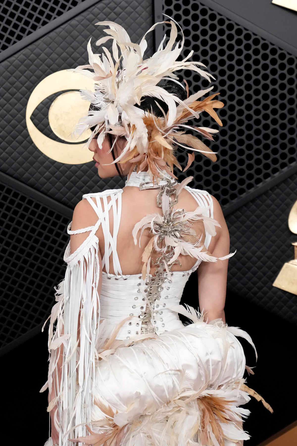 Person wearing an elaborate white feathered costume and headpiece, seen from the back, on a black and gold decorated red carpet event backdrop with geometric patterns.