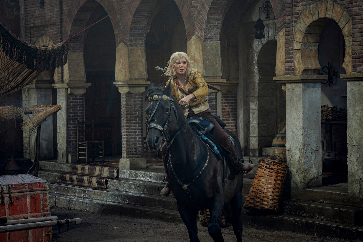 A person with blonde hair riding a black horse through a rustic, arched stone courtyard, dressed in medieval-style clothes, with a tense and adventurous atmosphere.