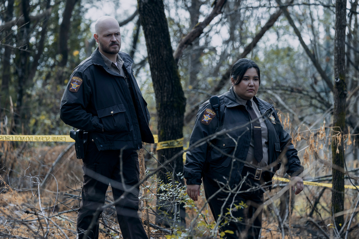 Two uniformed police officers stand in a wooded area with yellow caution tape, appearing to investigate a scene amid trees and dry underbrush.