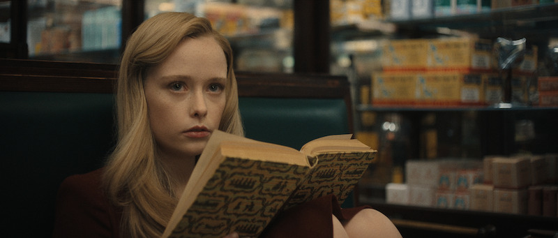 A young woman with long blonde hair sits in a booth, holding an open book and staring ahead with a serious expression in a store or diner setting.