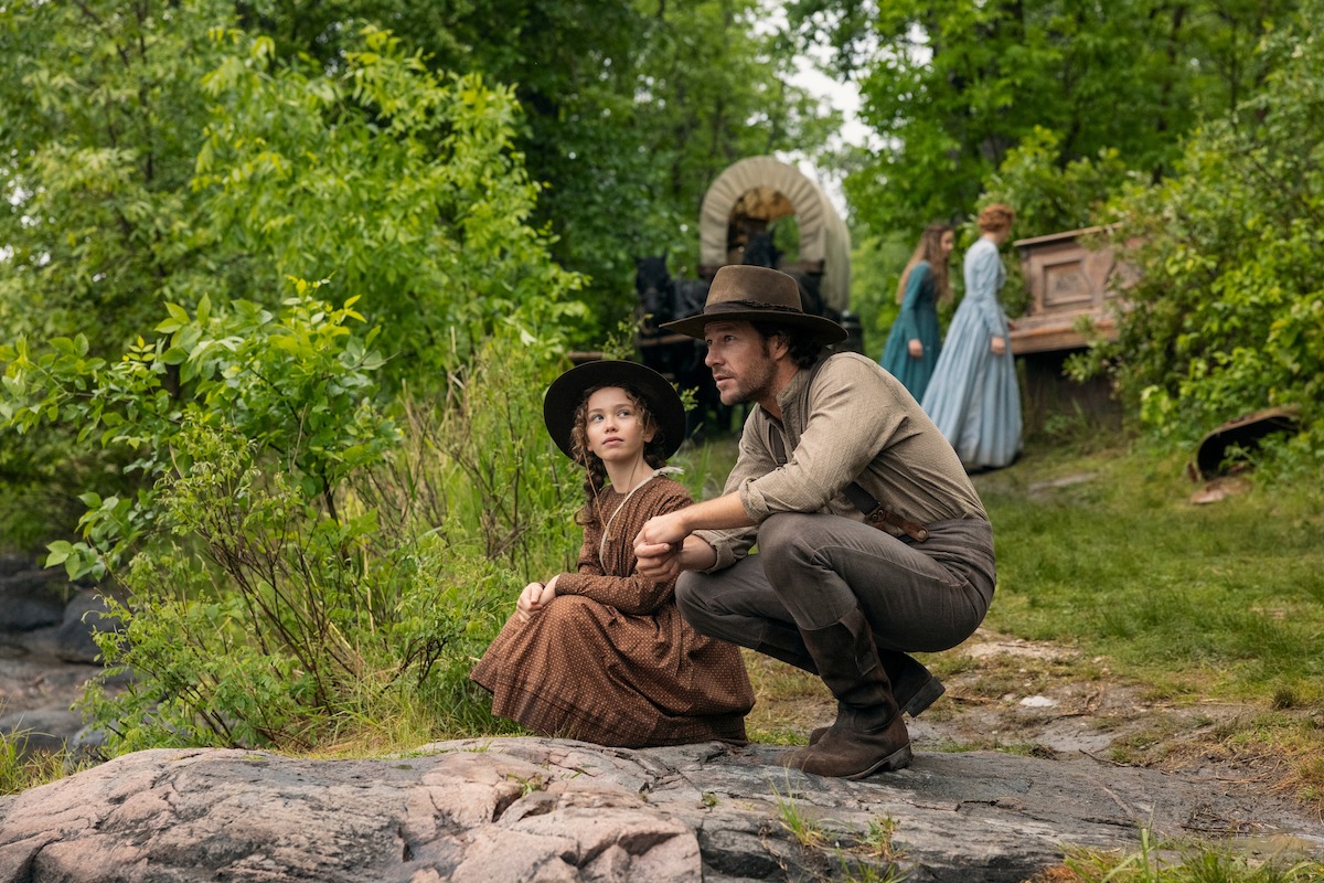 Man and young girl in pioneer-era clothing crouch together on rocky ground in a forest. Covered wagon and two women in period dresses stand in the background, suggesting a 19th-century frontier or pioneer setting.