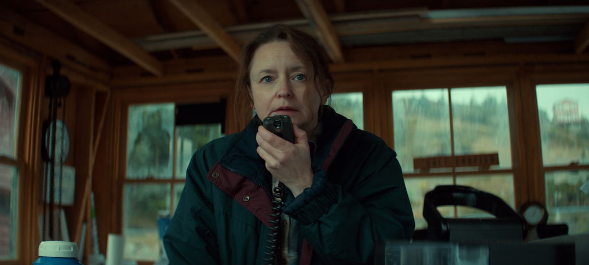 A woman in a green jacket speaks into a handheld radio inside a wooden cabin with large windows, surrounded by communication equipment and natural outdoor light.
