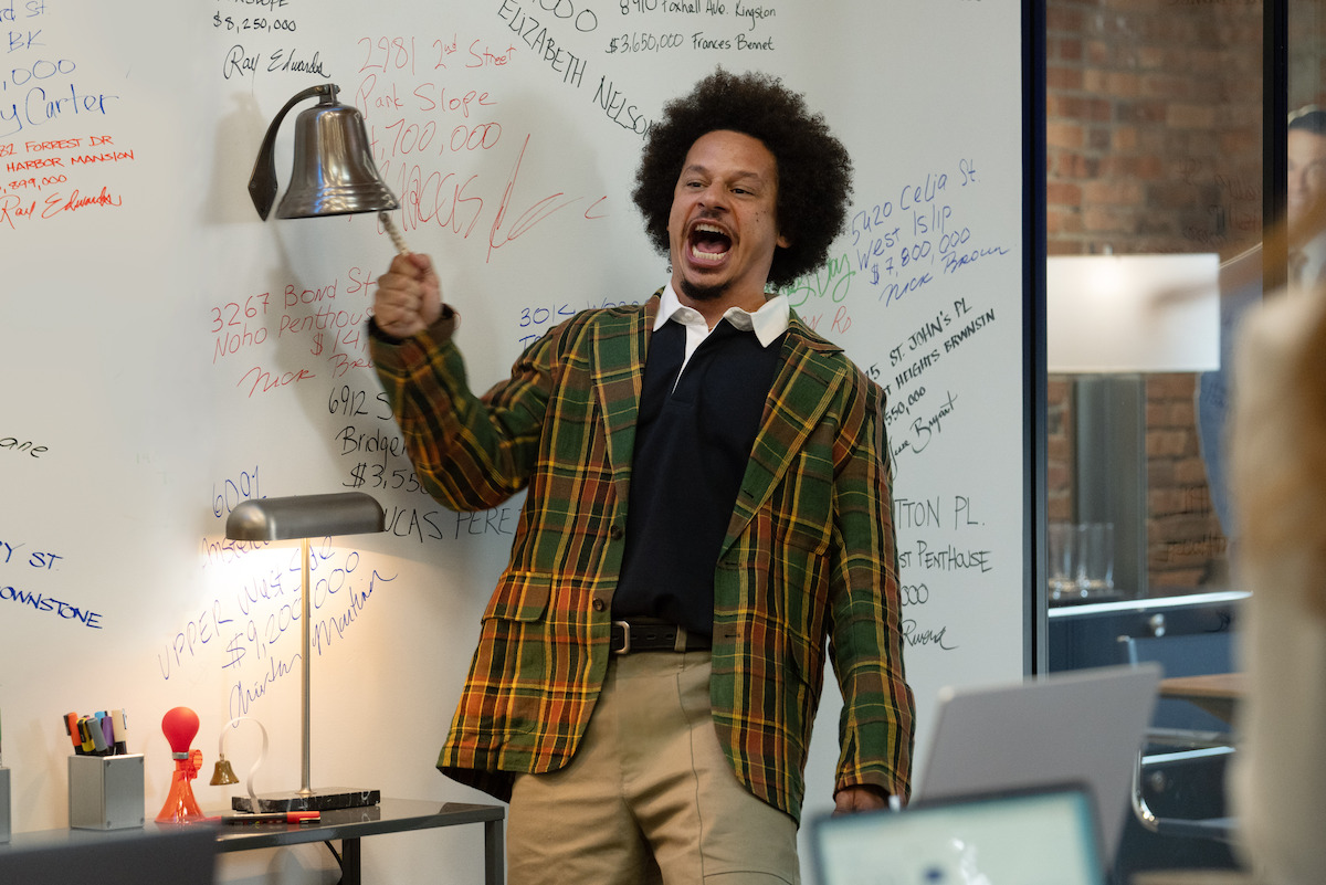 Man in a plaid jacket enthusiastically ringing a bell in a modern office, standing by a whiteboard covered in colorful writing. Desk with lamp and office supplies visible, with colleagues working in the foreground.