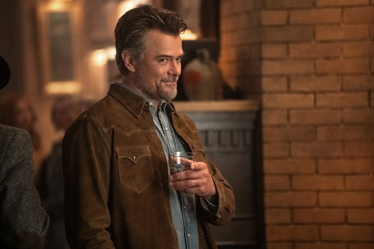 A man with gray hair and beard wearing a brown suede jacket stands indoors by a brick wall, holding a glass and smiling in a warmly lit, cozy setting that appears to be a social gathering or party.