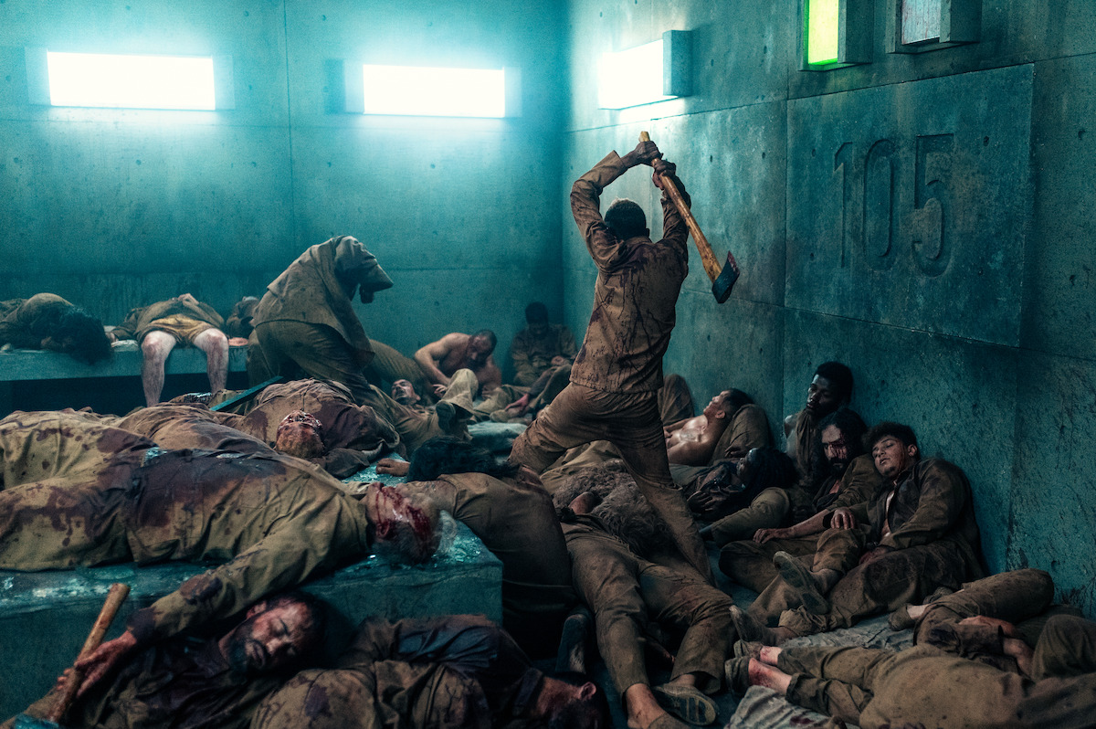 In an image from the film ‘The Platform 2,’ in a concrete cell with the numbers 105 etched into the wall, bloody bodies in khaki uniforms are spread across the floor while a man swings an ax above them.