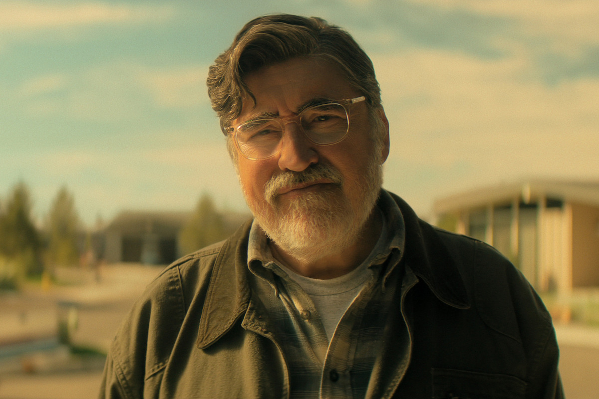 Alfred Molina as Sam, with glasses and gray hair standing outdoors, wearing a jacket and plaid shirt on a sunny day, with modern buildings and trees blurred in the background.