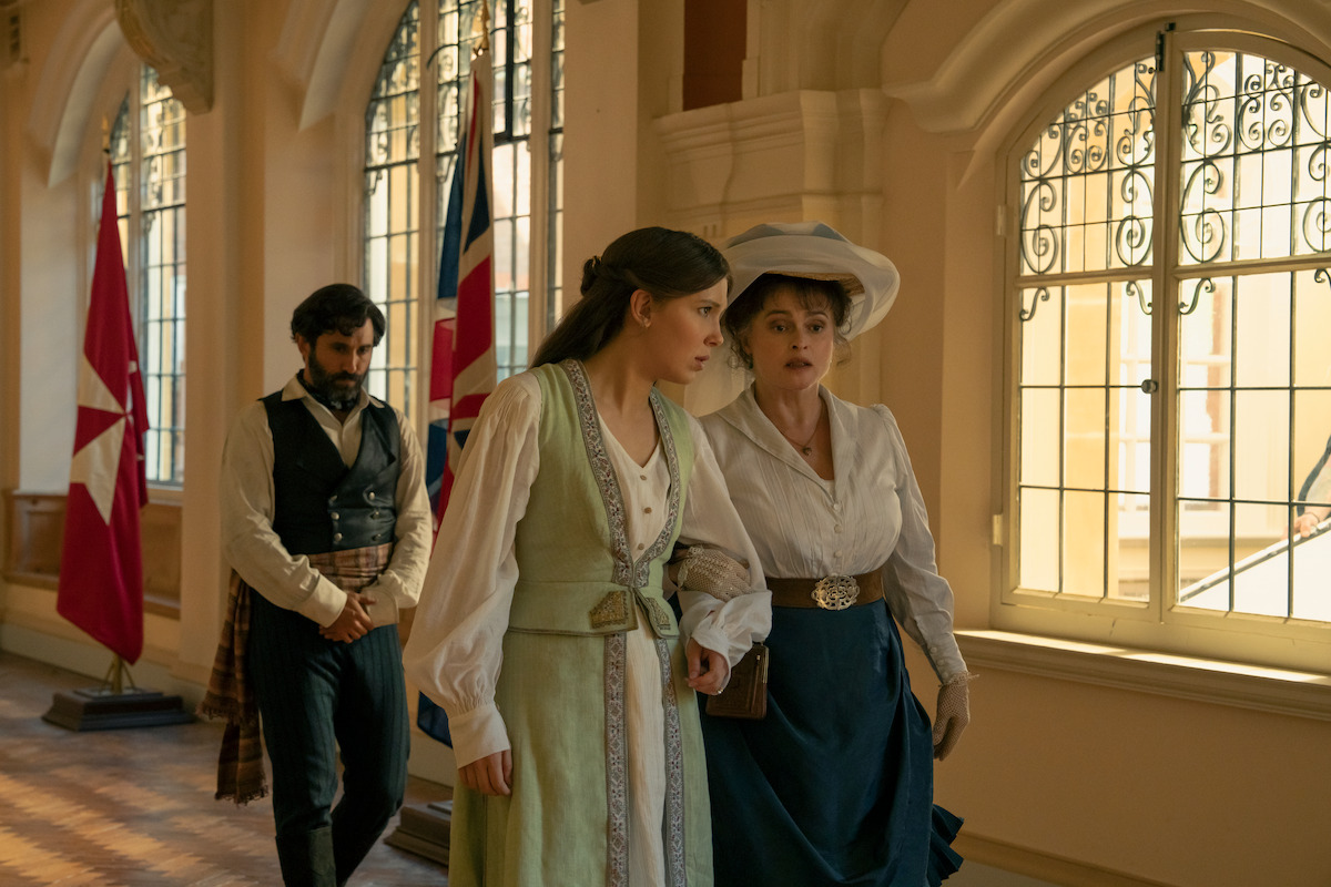 Three people in period costumes walk in a sunlit hallway with large arched windows and flags, suggesting a historical or formal setting. Two women walk arm-in-arm while a man follows behind.