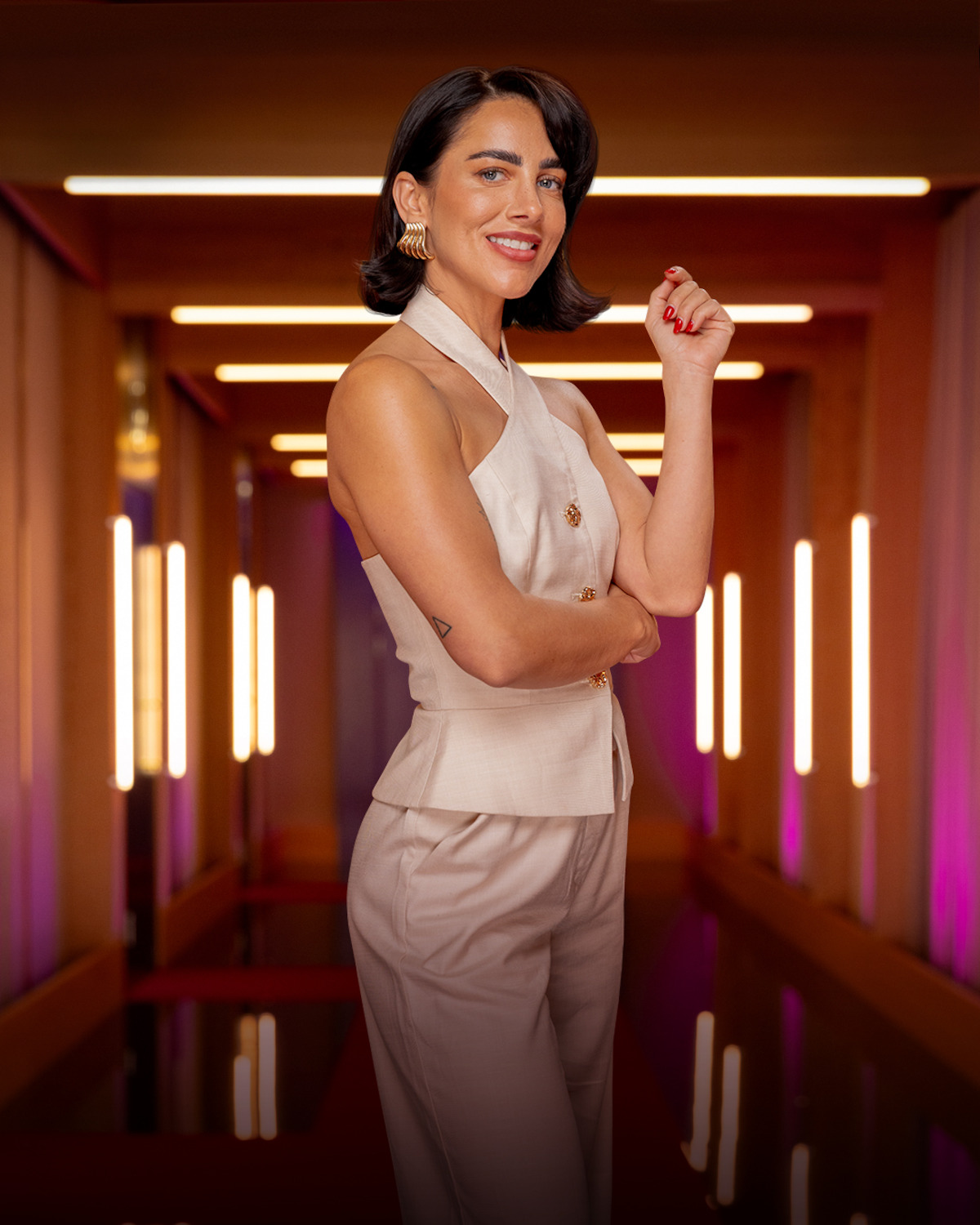 Woman in a sleeveless beige outfit stands confidently in a stylishly lit hallway with warm ambient lights and modern decor, smiling and posing with one arm raised, creating an elegant and fashionable atmosphere.