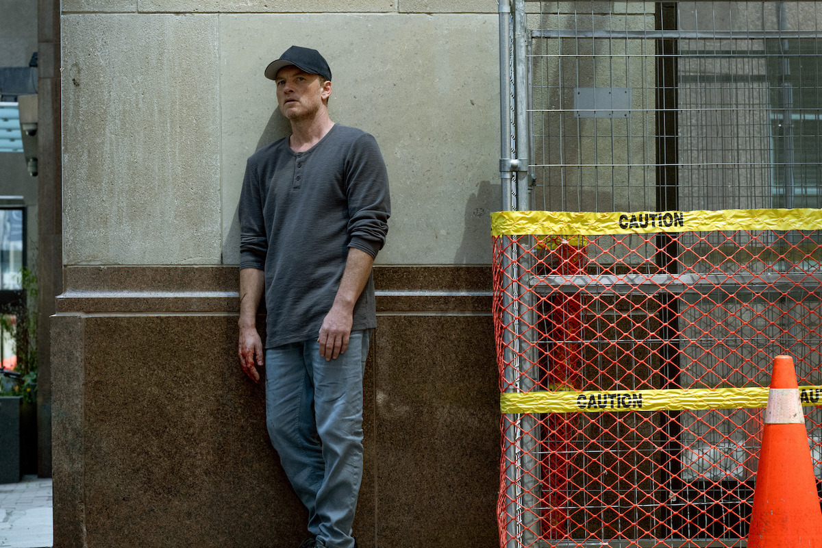 A man in casual clothes and a cap stands against a building wall near a construction site marked with orange netting, yellow caution tape, and a traffic cone in an urban setting.
