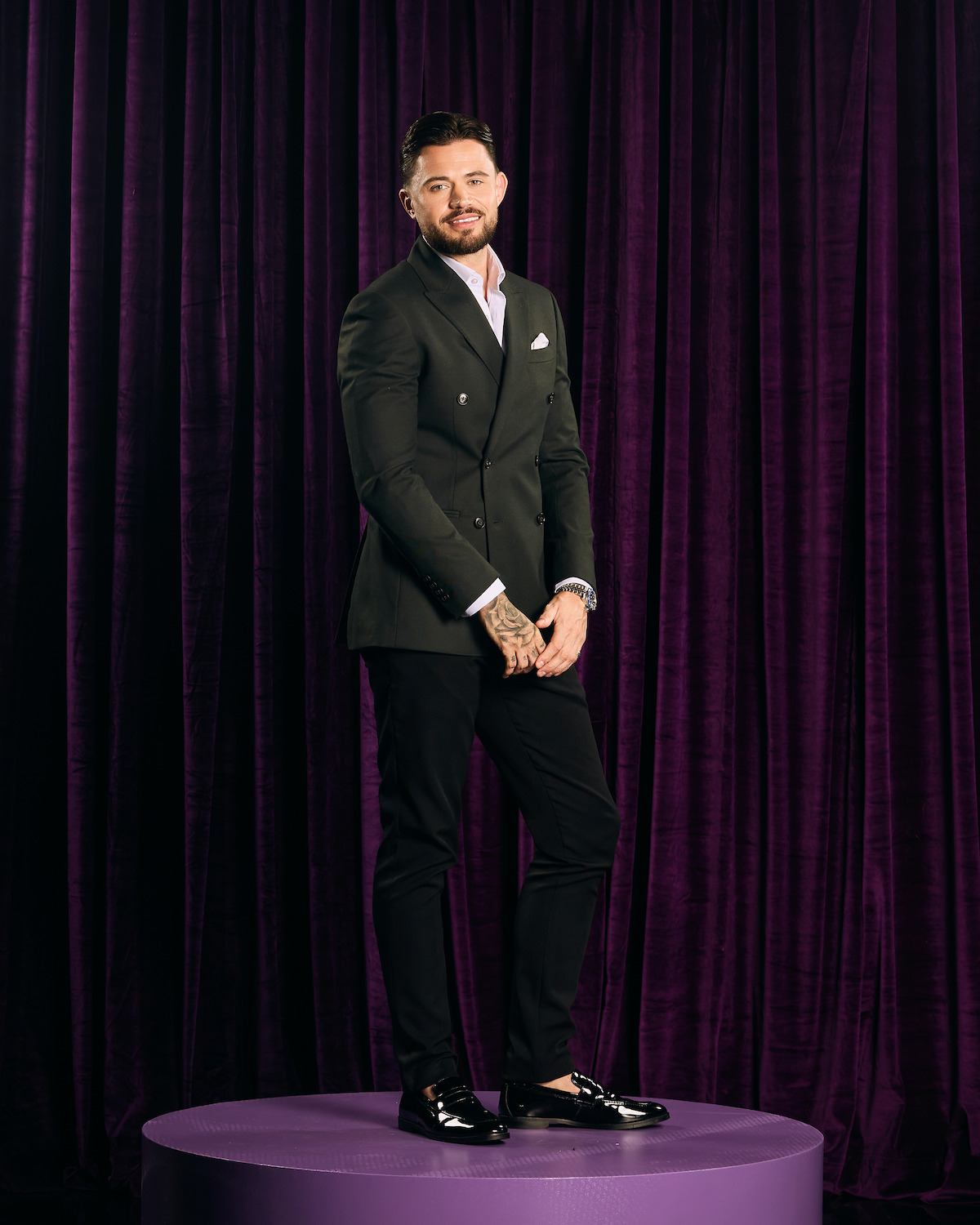 A well-dressed man with tattoos stands on a round purple platform in front of dark purple curtains, wearing a suit jacket, dress shirt, and loafers in a formal indoor setting.