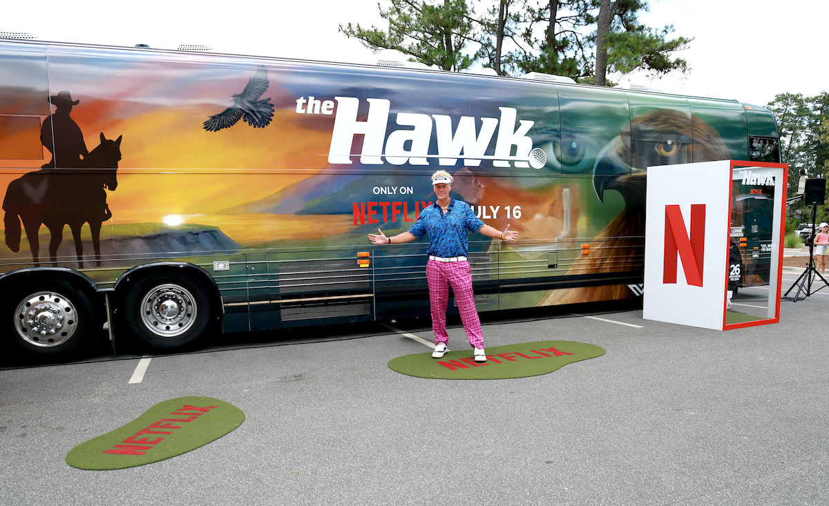 Person posing in front of a tour bus with "the Hawk" and Netflix branding, outdoors in a parking area with pine trees, promotional setup includes large Netflix logo and branded green mats on the ground.