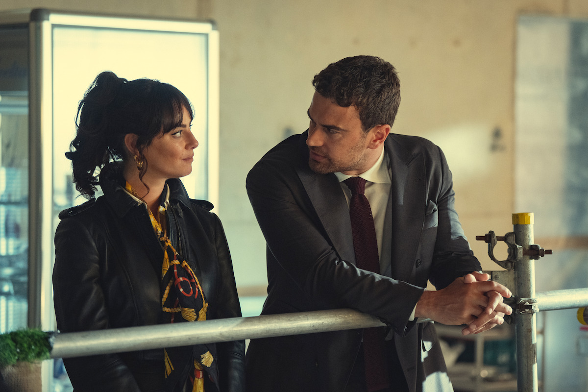 A woman in a leather jacket and a man in a suit stand together indoors, leaning on a railing, looking at each other in conversation, with industrial and business-like surroundings visible in the background.