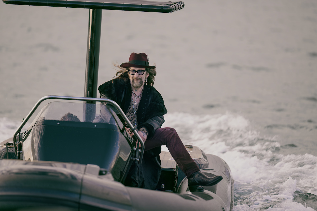 Paul Kaye wears an eccentric outfit with eye glasses and appears to be driving a small motorized raft boat.