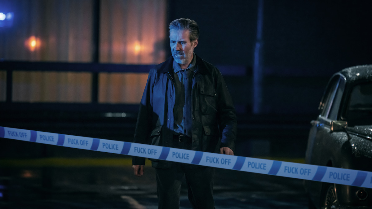 A serious man stands behind blue police tapeat night, next to a car and in front of a building with lit windows. The scene appears tense and dramatic, with wet pavement reflecting lights.