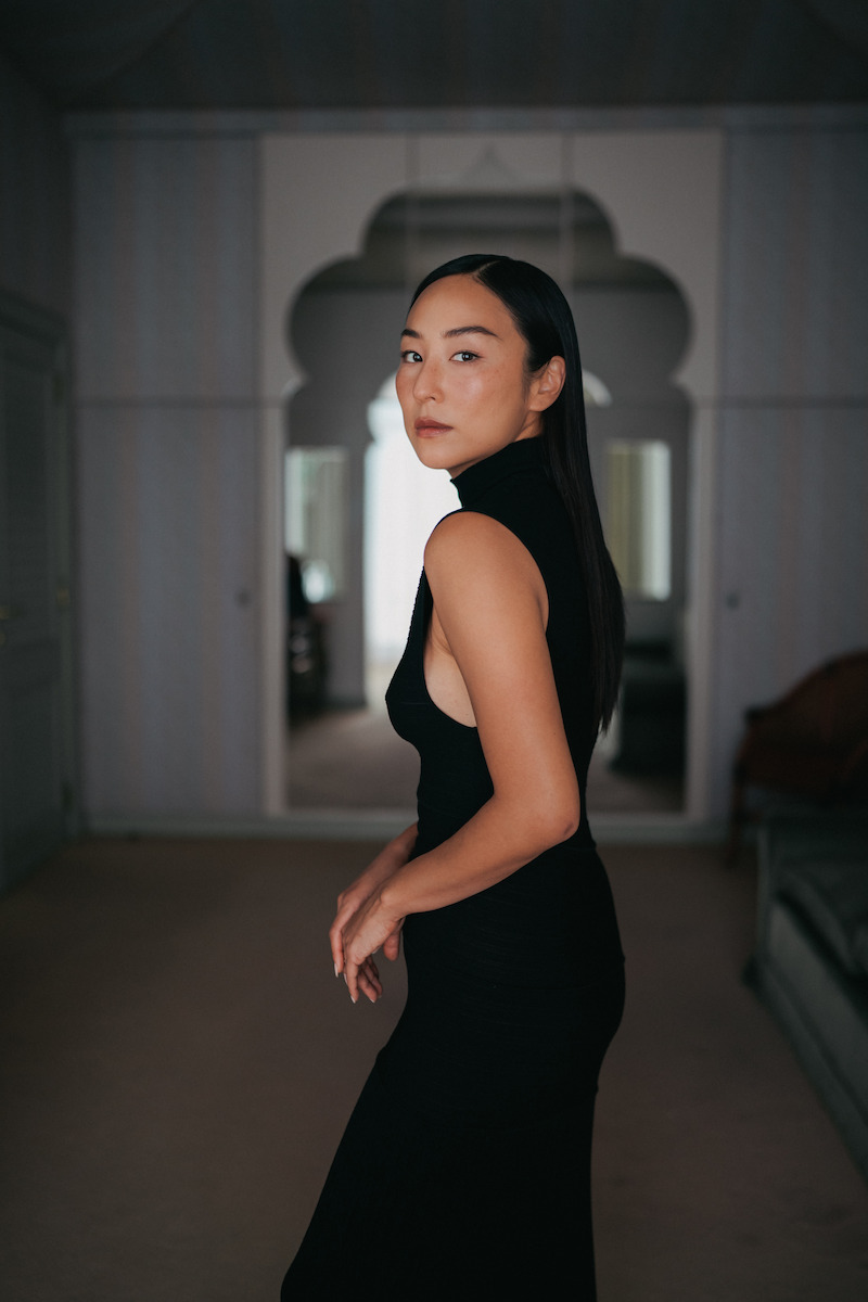 Greta Lee in a black sleeveless dress poses in a hotel room. 