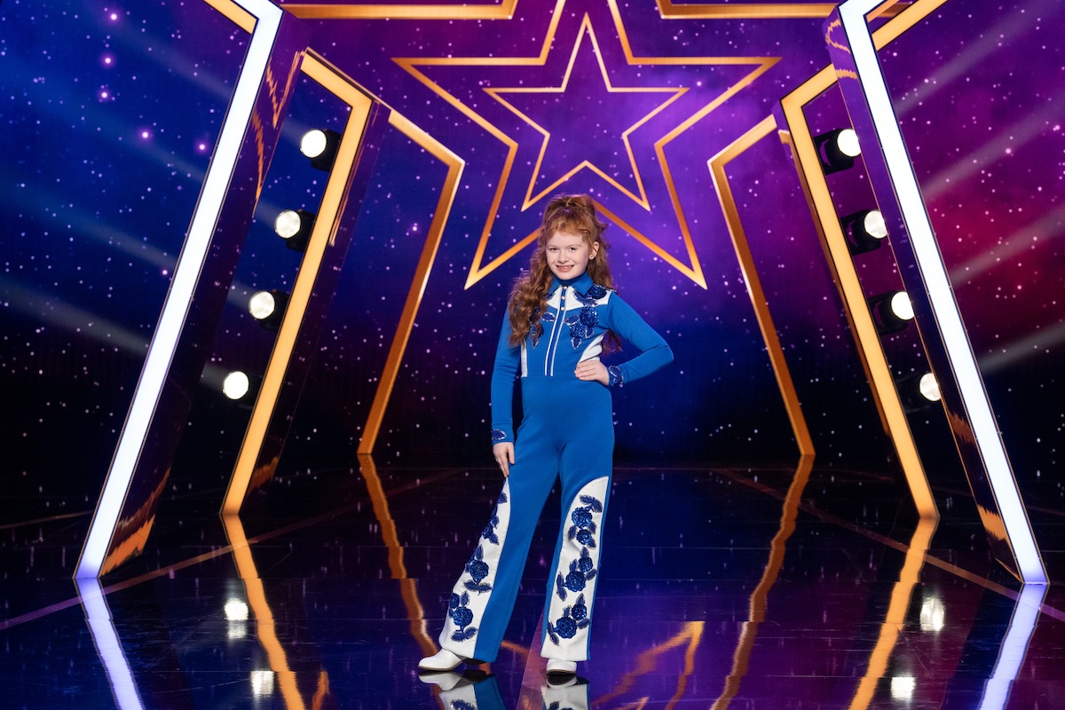 Blair Kudelka, con un mono azul, se encuentra con confianza en un escenario brillante con un gran fondo de estrellas, rodeado de luces brillantes y un fondo colorido de temática cósmica.