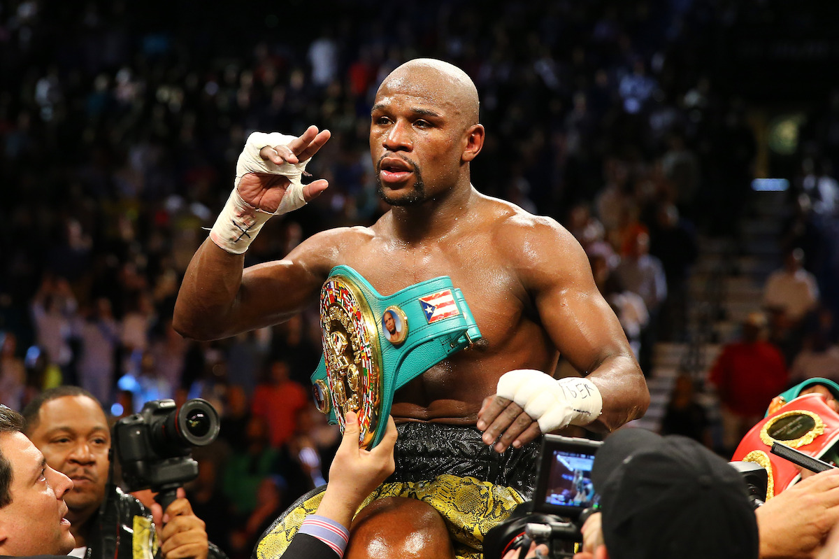 Floyd Mayweather Jr., sweaty and victorious, holding a championship belt amid cheering crowd and photographers after a match, under bright arena lights.