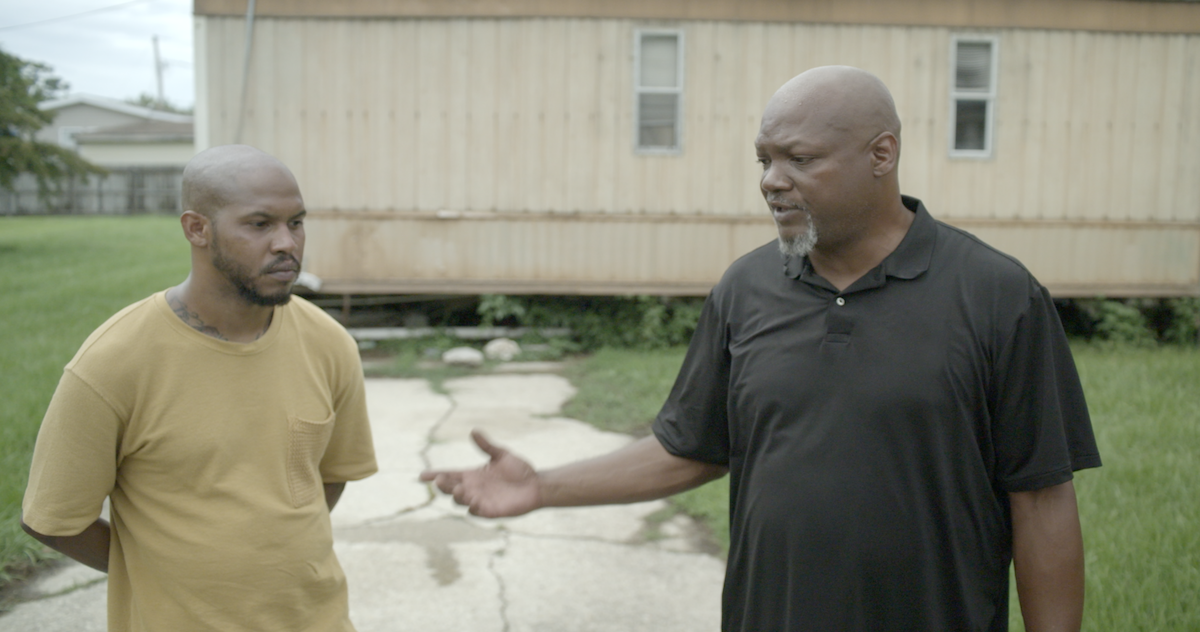 Two men solemnly talking in front of a mobile home.