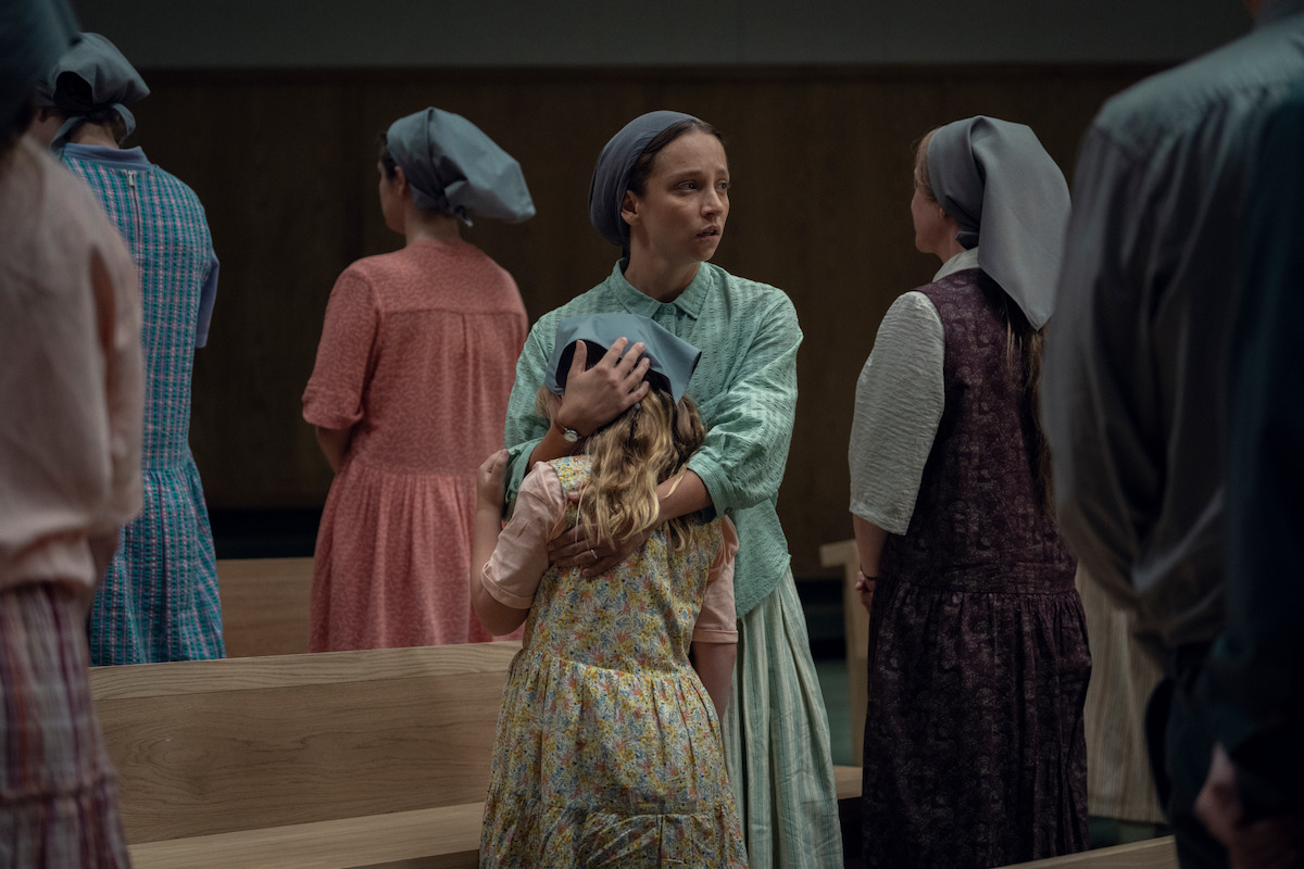 A woman in plain clothing and headscarf embraces a child in a modest, wood-paneled room among other similarly dressed women, suggesting a communal or religious gathering.