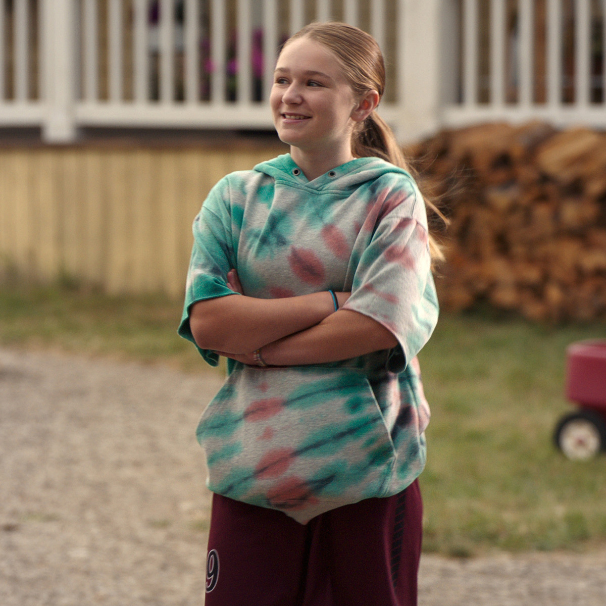 Smiling girl in a tie-dye hoodie stands with arms crossed outside near a wooden porch, stacked firewood, and a red wagon in the background.
