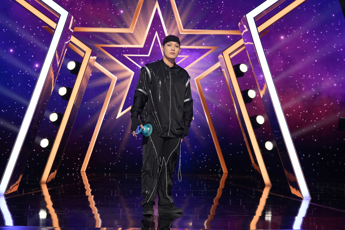 Chihhan Chao in a black tracksuit holding a diabolo on a shiny stage with star-shaped purple and gold lights and spotlights in the background, creating a vibrant talent show atmosphere.
