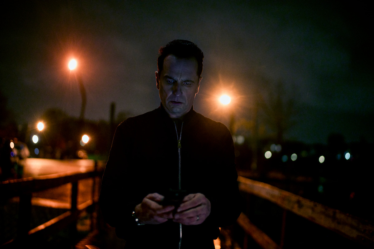 A man stands on a wooden walkway at night, illuminated by streetlights, looking at his phone. The environment is dark and moody, with a somber, contemplative atmosphere.