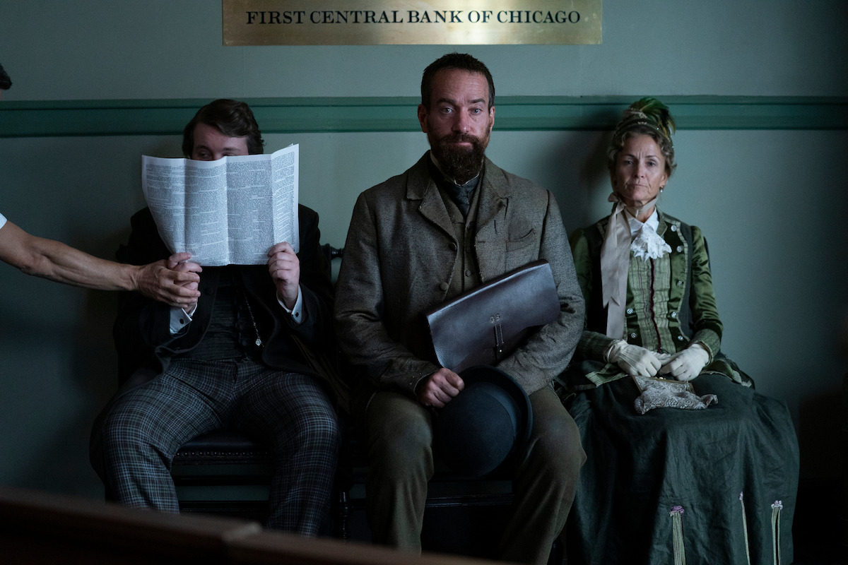Three people in period clothing sit on a bench inside a vintage bank lobby, one reading a newspaper, another holding a hat and folder, and a third woman in a green dress, with a serious and tense atmosphere.