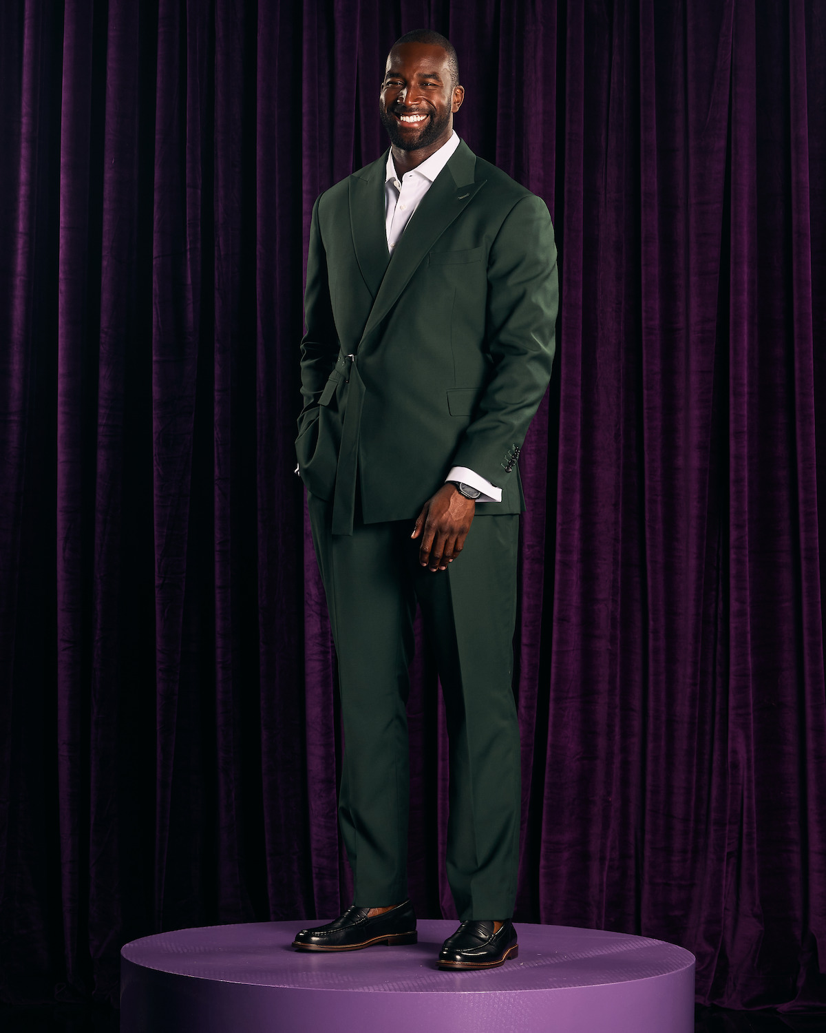 A man in a dark green suit stands on a round purple platform in front of deep purple curtains, smiling, in a formal indoor portrait setting.